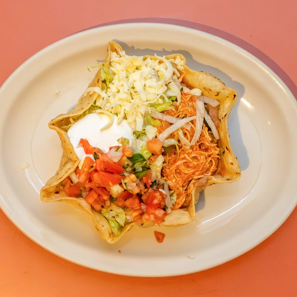 A taco salad in a crispy tortilla bowl, topped with shredded chicken, lettuce, cheese, pico de gallo, and sour cream.