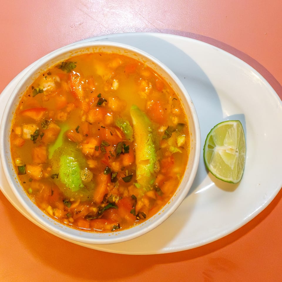 A vibrant orange soup with avocado slices and herbs, served with a lime wedge.