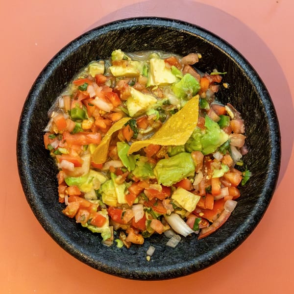 A black bowl filled with chunky guacamole, pico de gallo, and two tortilla chips on a peach surface.